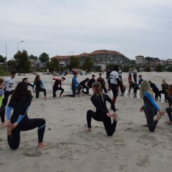 Zdjęcie grupy uczniów ćwiczących na plaży. Wszyscy ubrani są w kombinezony. Widać, że rozciągają mięśnie czworogłowe w klęku. Być może po rozgrzewce pójdą pływać.