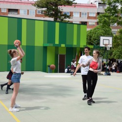 Zdjęcie uczniów na podwórku przed zielonym budynkiem. Na dwóch boiskach grają w koszykówkę.  Inni uczniowie odpoczywają przy drzewach znajdujących się po prawej stronie za boiskiem.