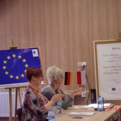 obrazek w kolorze przedstawiający na pierwszym planie dwie kobiety, w tle tablica unii europejskiej i tablica z tytułem projektu