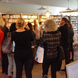 Na kolorowym obrazku widać grupę osób ( widok z tyłu) w sali bibliotecznej, przed nimi mężczyzna w garniturze i krawacie