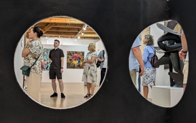 Oprowadzanie kuratorskie w Muzeum w Koszalinie Zdjęcie przedstawia ludzi zwiedzających wystawę, widocznych przez dwa duże otwory umieszczone na czarnym tle.