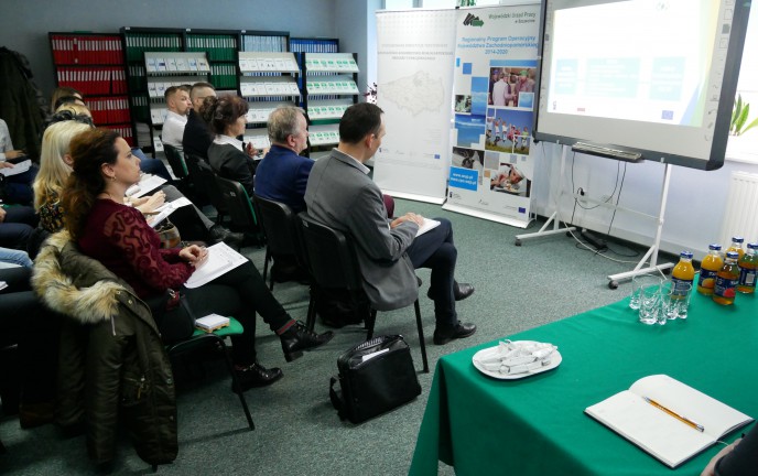 grupa osób siedzi na krzesłach w dwóch rzędach, oglądająca slajdy na ekranie