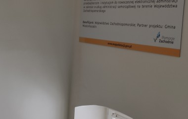 obrazek w kolorze przedstawiający tablicę informacyjną projektu zawieszoną nad oknem, wewnątrz budynku Urzędu Miejskiego w Koszalinie