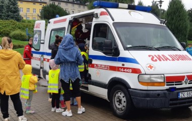 obrazek w kolorze przedstawia grupkę dzieci z dwiema opiekunkami oraz ambulans pogotowia ratunkowego 