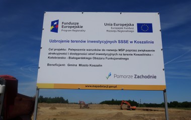 Kolorowy obrazek przedstawiający fragment terenu Strefy Inwestycyjnej w Koszalinie z umiejscowioną w centrum tablicą informacyjną. 