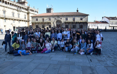 Zdjęcie grupy uczniów wraz z ich opiekunami. Znajdują się oni na placu w mieście Vigo w Hiszpanii. Jest słonecznie. Wszyscy są zadowoleni.