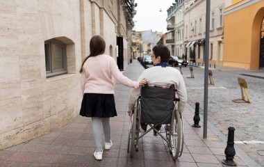 Na zdjęciu idzie dziewczynka i prowadzi wózek inwalidzki z osobą niepełnosprawną