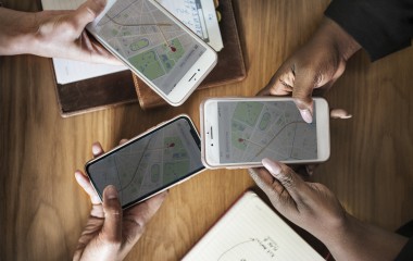 Na zdjęciu znajdują się trzy ręce trzymające w dłoniach telefony komórkowe z odpalonymi mapami online. 