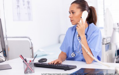 Pani pielęgniarka rozmawiająca przez telefon. Siedzi za biurkiem. Przed sobą ma monitor