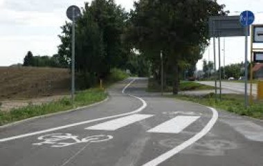 Fragment ścieżki rowerowej z przejściem dla pieszych oraz z przystankiem autobusowym z prawej strony, ciągnącej się dalej wzdłuż kłębistych ciemnozielonych drzew.