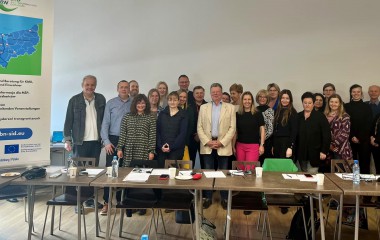 obrazek przedstawia w kolorze od lewej stojący pionowy baner projekktu SID, na ustawione ławy, na których rozłozone są miedzy innymi długopisy, kartki,za ławami stoi liczna grupa osób - uczestników projektu 